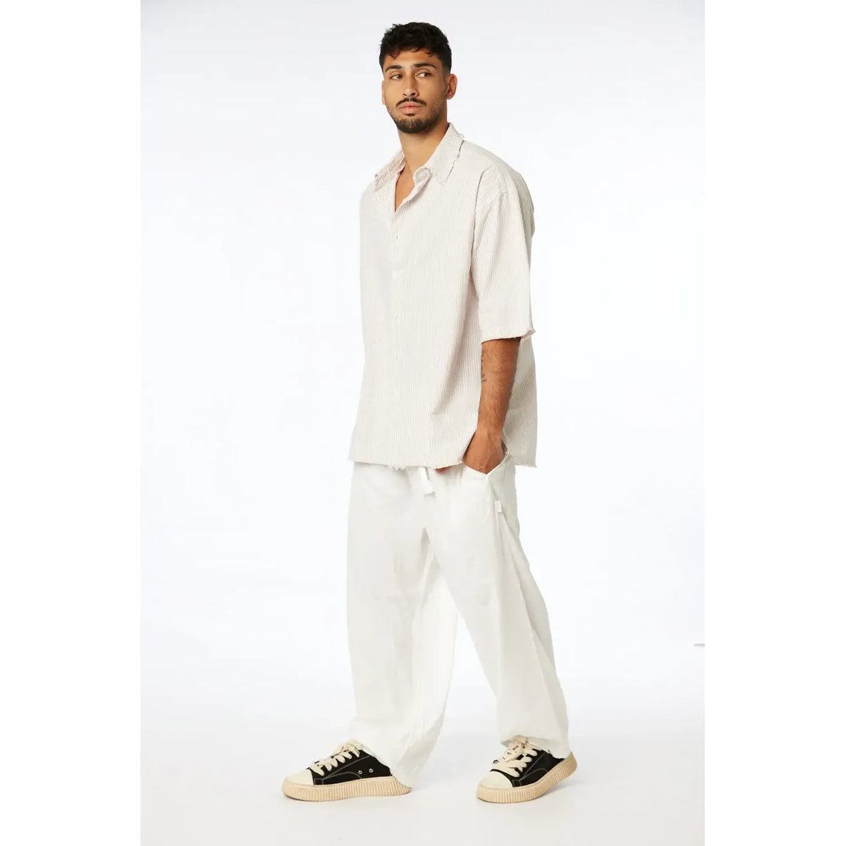 Man wearing a Striped Linen Short Sleeve Button-Up Shirt in Stone, white linen pants, and black and white sneakers