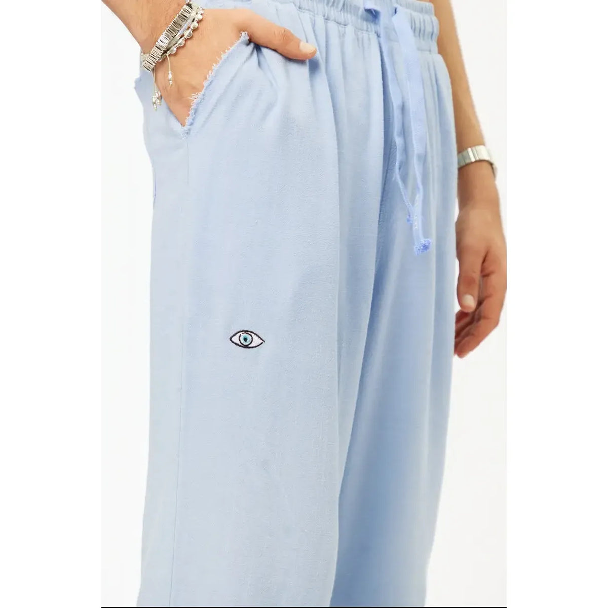 Close up of sky blue SOFT SL Linen Pants with a small embroidered eye on the left leg and a hand in the pocket