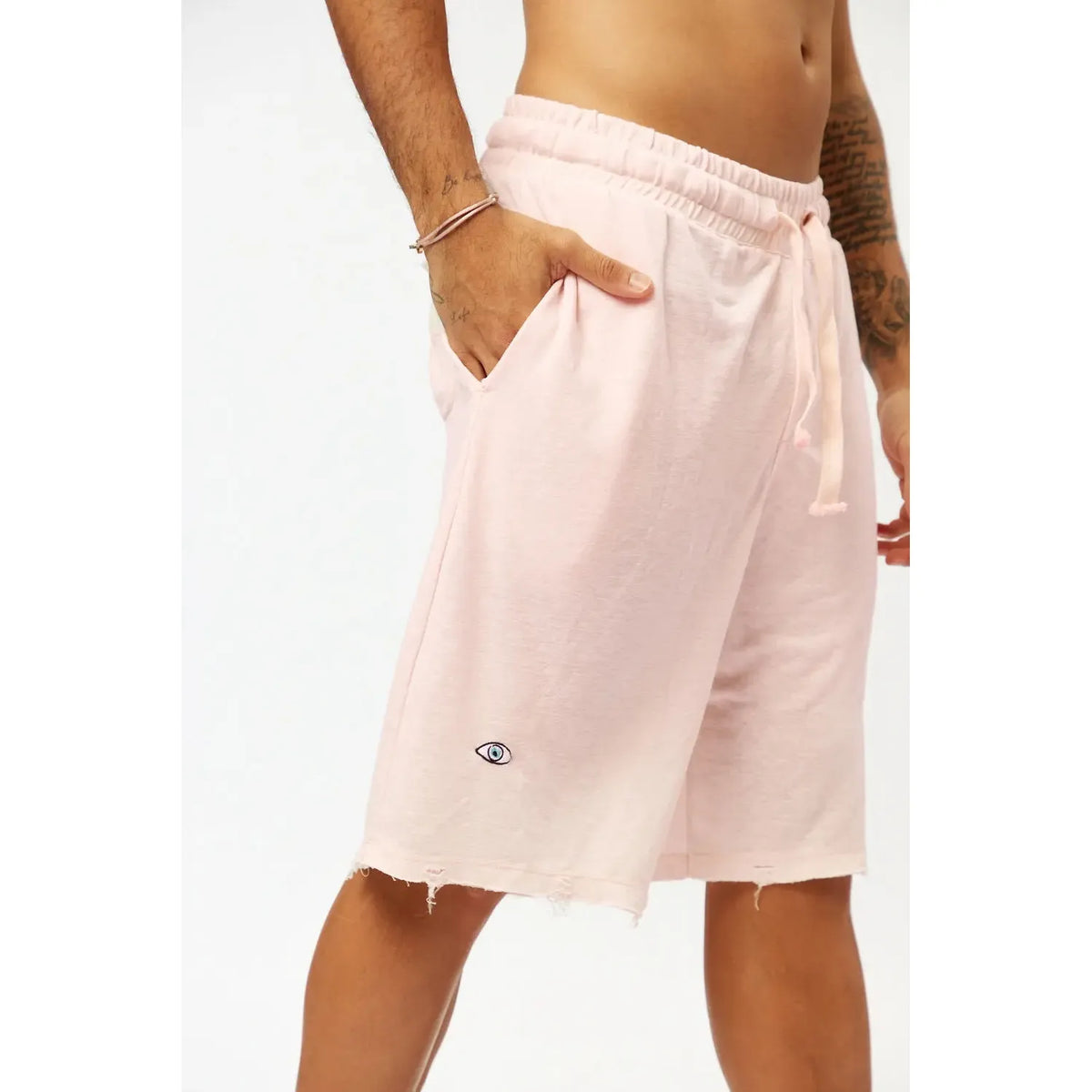 Base Shorts in baby pink with a small eye logo on the left leg, worn by a person with their hand in the pocket