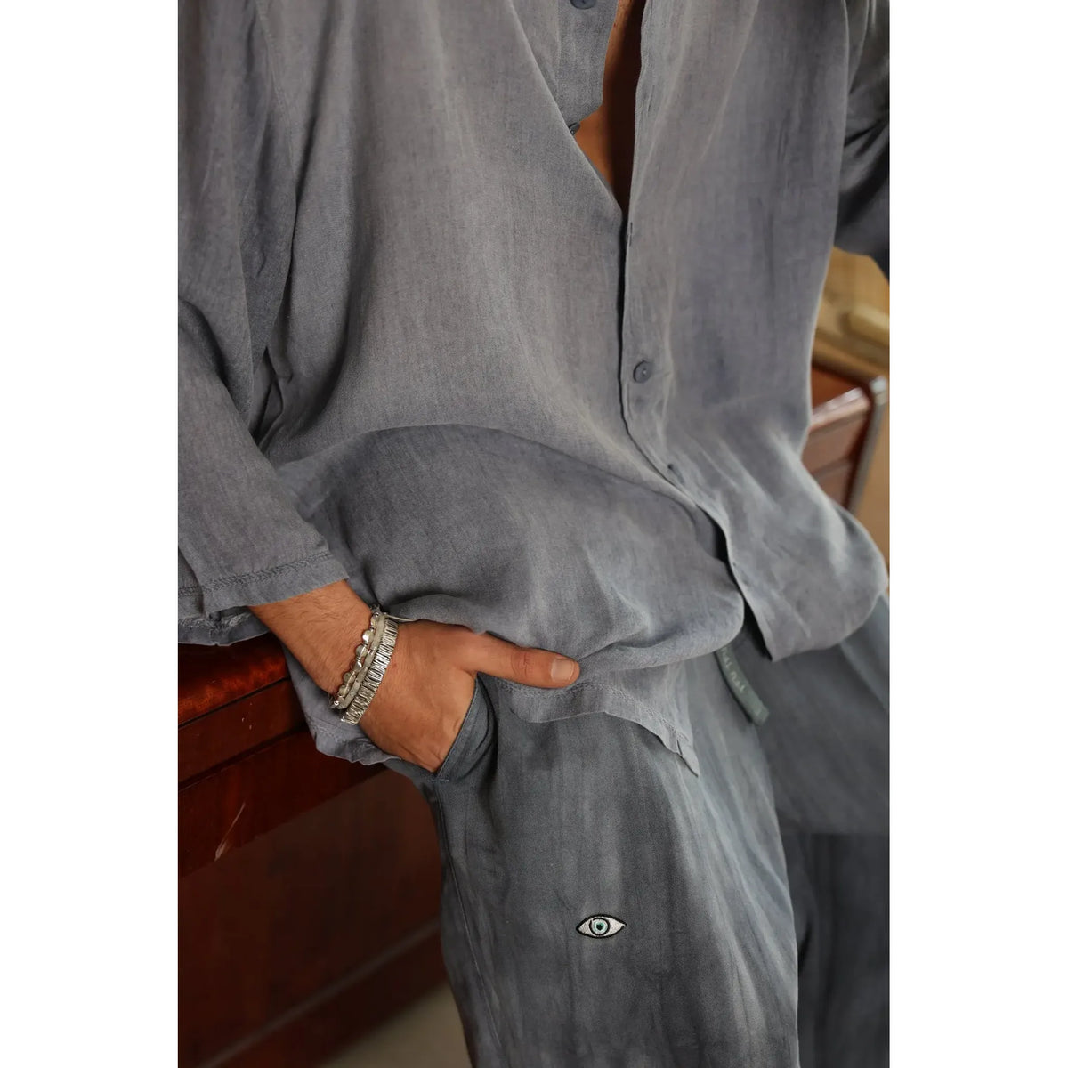 Man wearing an open VENICE Button-Up Shirt in ocean blue, with a hand in his pocket, showing bracelets and an eye embroidery on the pants