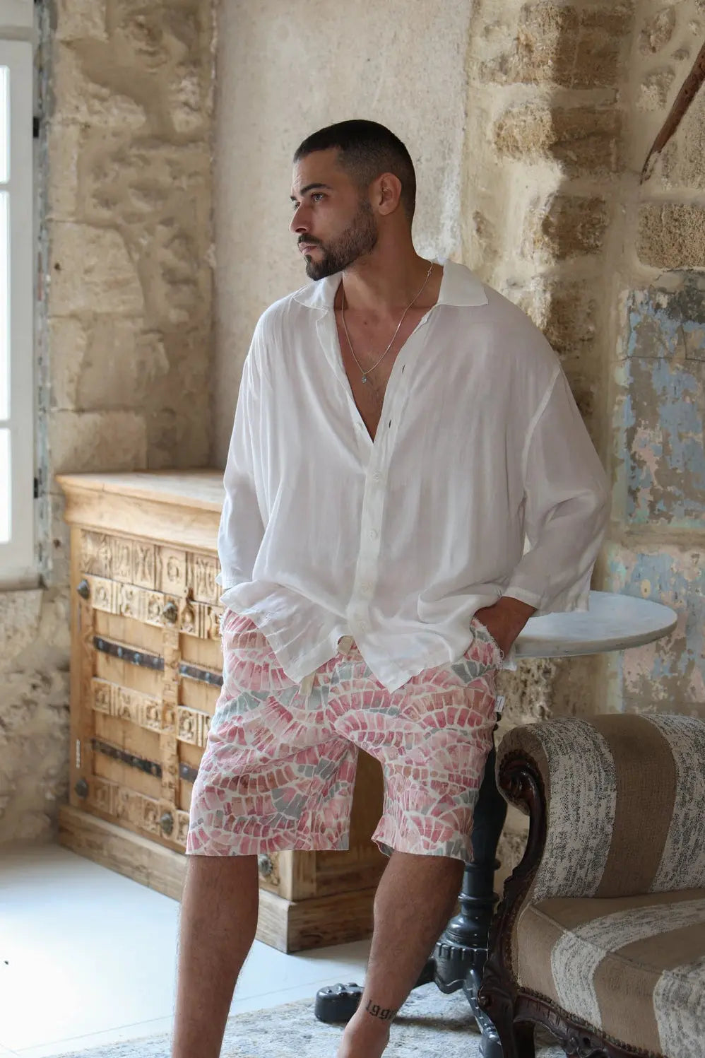 Man wearing a white shirt and patterned shorts in a rustic room.
