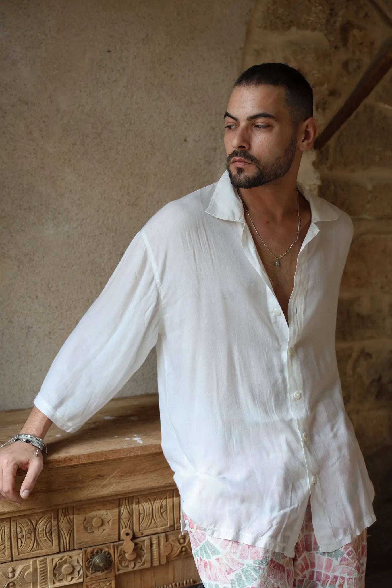 Man wearing a white VENICE Button-Up Shirt and patterned shorts, leaning on a carved wooden chest
