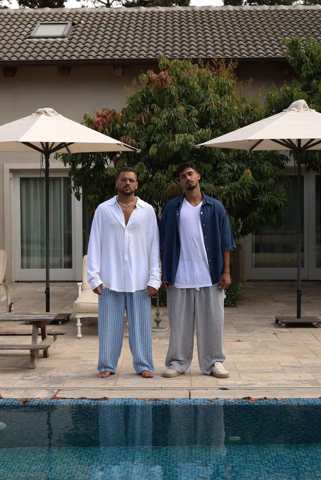 Two men in Pure Oversized Linen Pants, one in blue and white stripes, the other in grey and white stripes, by a pool