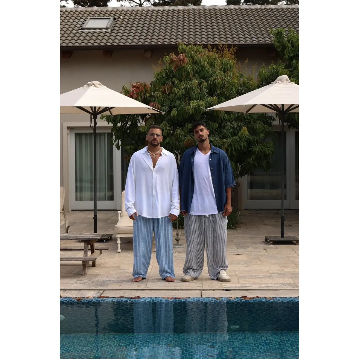 Two men in Pure Oversized Linen Pants, one in blue and white stripes, the other in grey and white stripes, by a pool