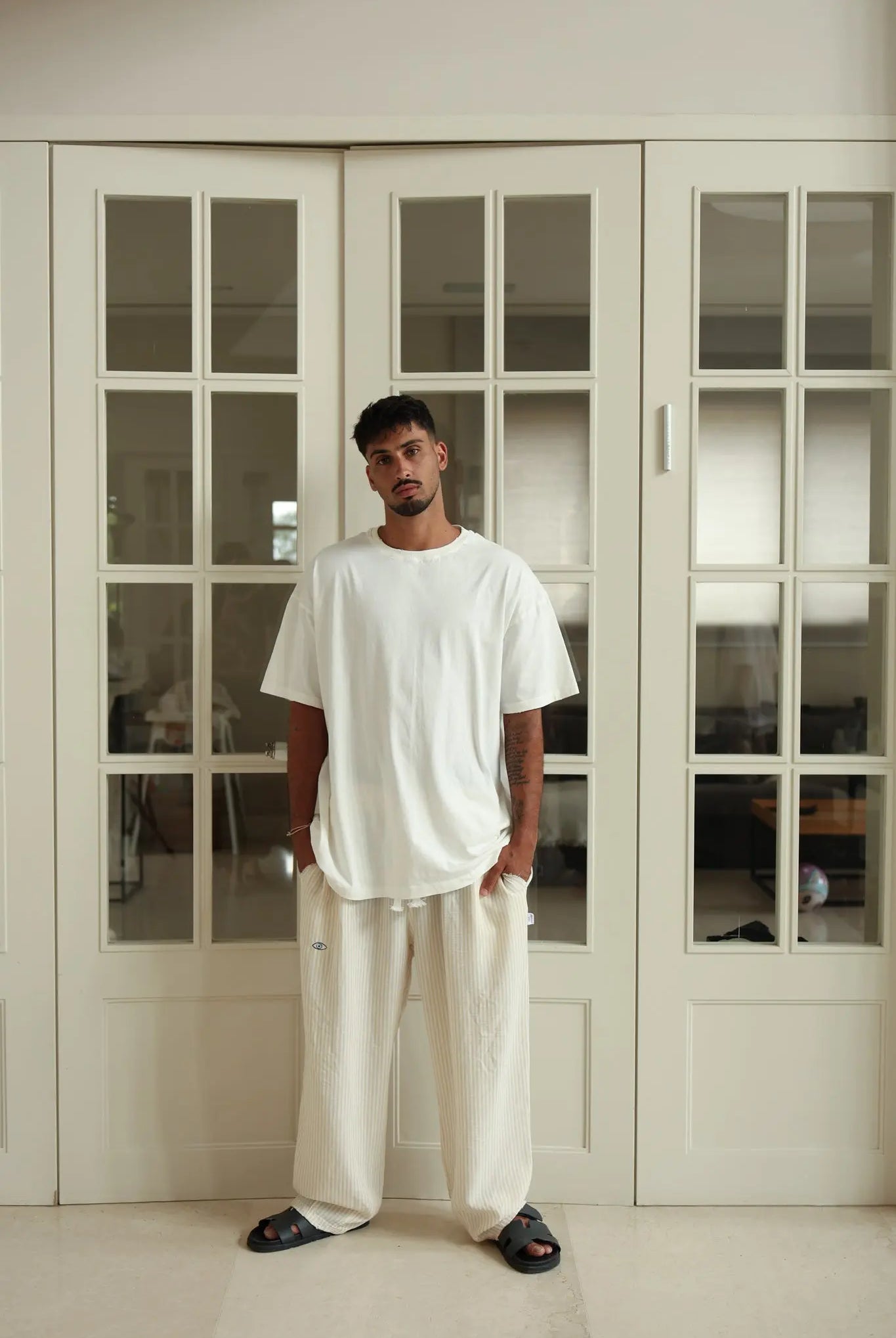 Man wearing a white t-shirt and pants standing in a room with large glass doors.