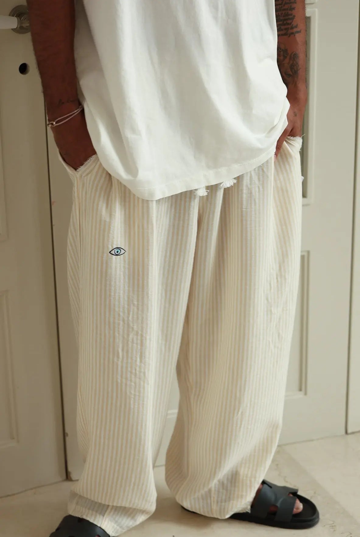 Pure Oversized Linen Pants in yellow and white stripes with an eye graphic, worn by a person in black sandals