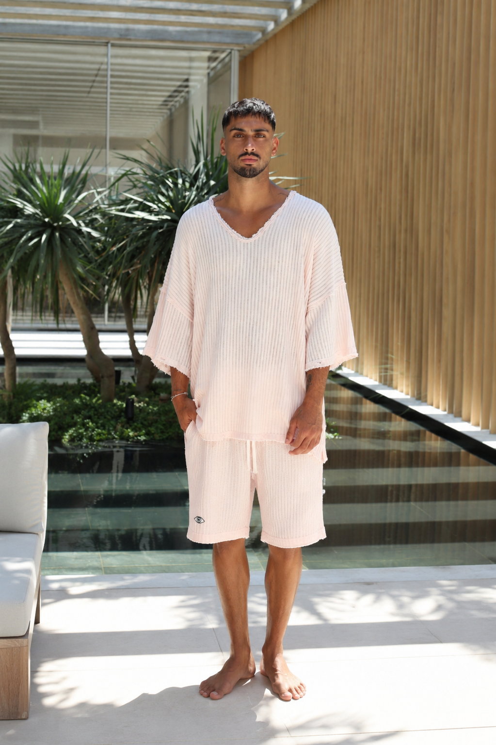 A man wearing a light pink ribbed lounge set consisting of a V-neck tunic top and matching shorts, standing barefoot in a modern outdoor patio setting with plants and a wooden slatted wall.