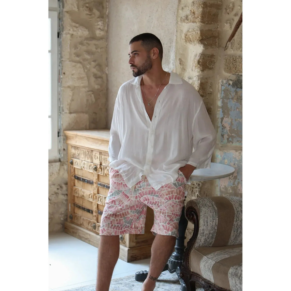 Man wearing a white shirt and patterned shorts in a rustic room.