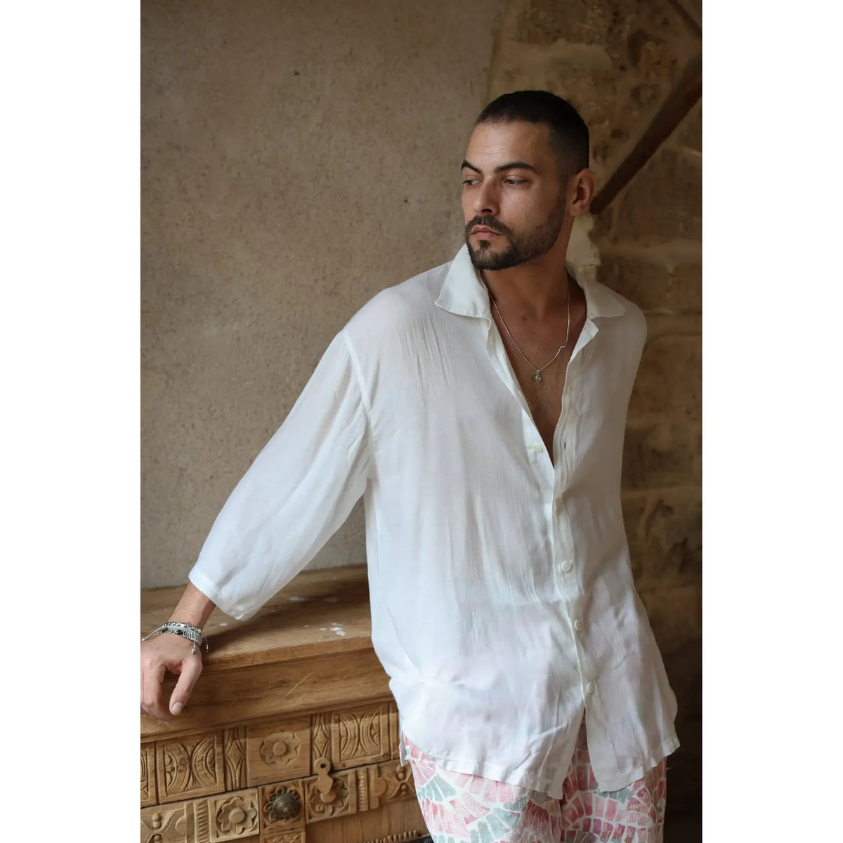 Man wearing a white VENICE Button-Up Shirt and patterned shorts, leaning on a carved wooden chest