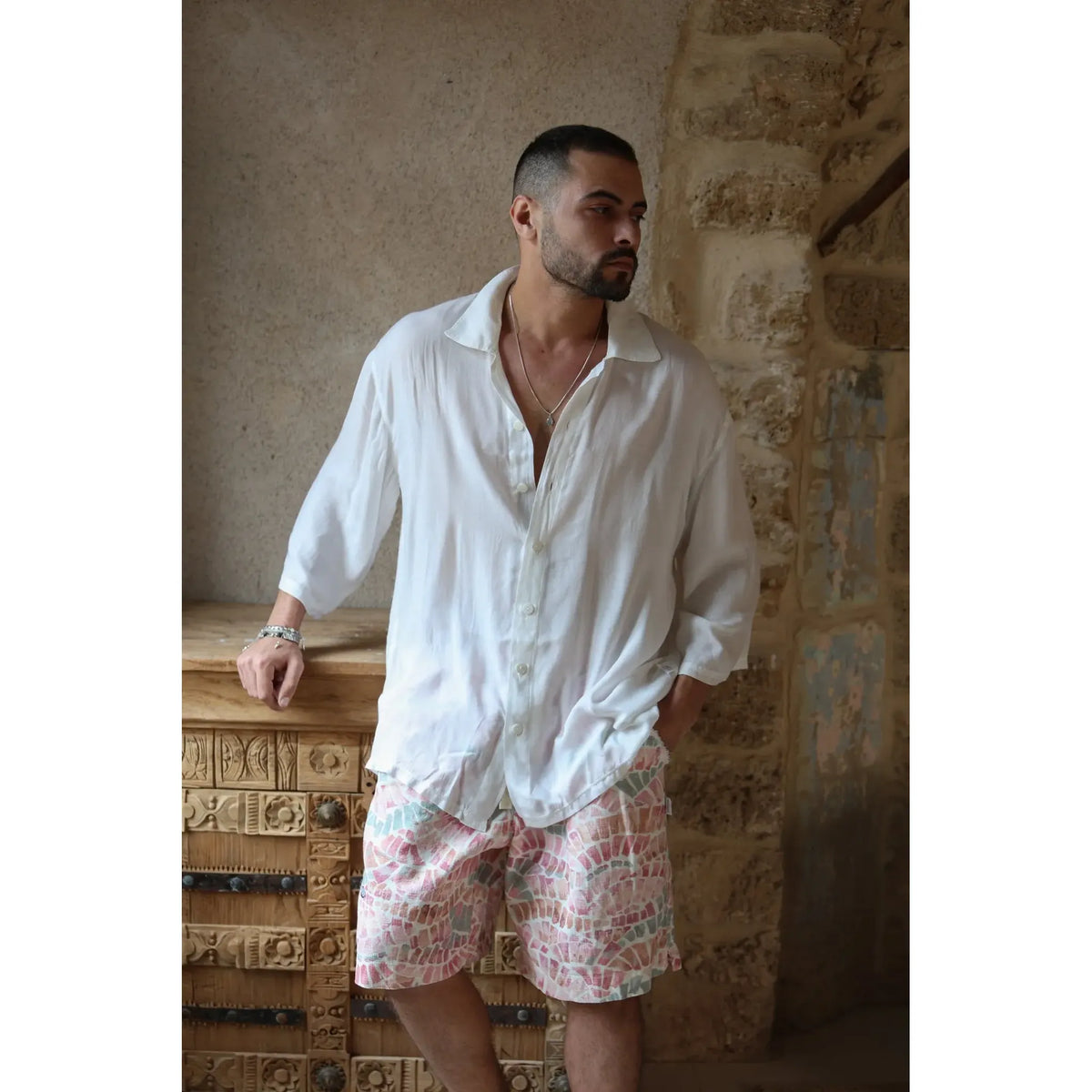 Man wearing a white VENICE Button-Up Shirt and patterned shorts, leaning on a carved wooden cabinet