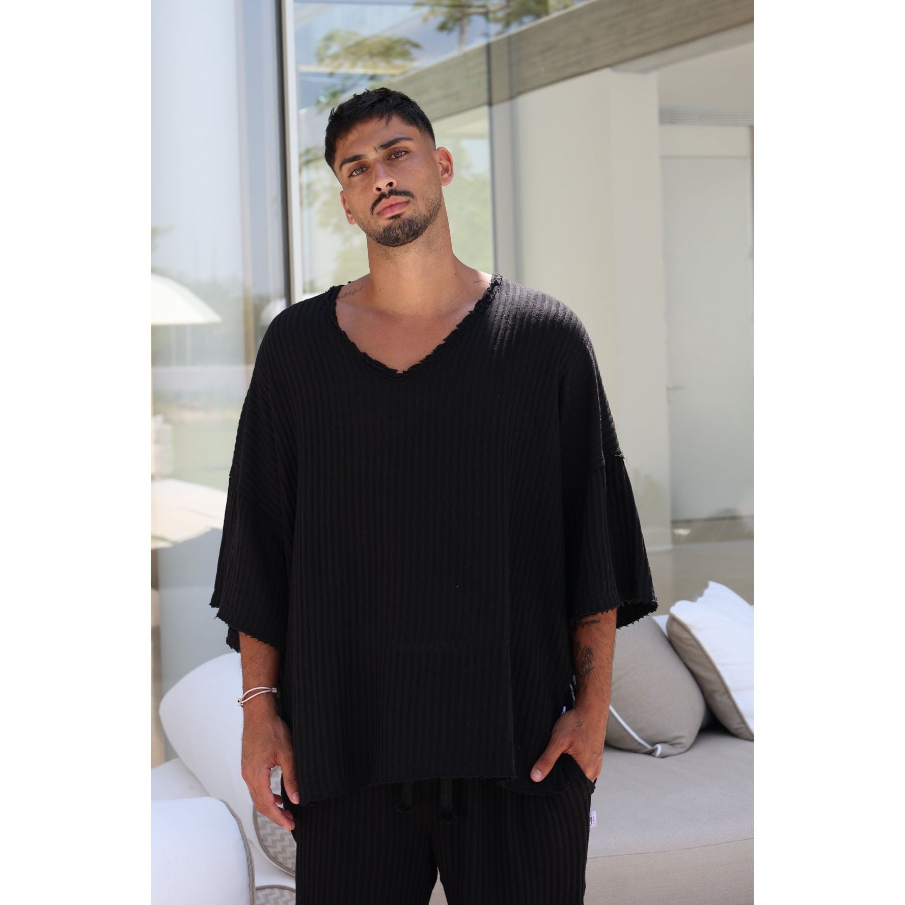 A man wearing a black ribbed V-neck tunic top with wide, tiered sleeves and matching black pants, standing in a modern, sunlit room with a white sofa in the background.