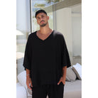 A man wearing a black ribbed V-neck tunic top with wide, tiered sleeves and matching black pants, standing in a modern, sunlit room with a white sofa in the background.