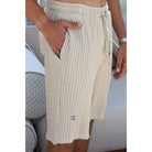 A close-up of a person wearing cream ribbed knit shorts with side pockets and a small blue and white eye logo near the hem, standing indoors near a light-colored sofa.
