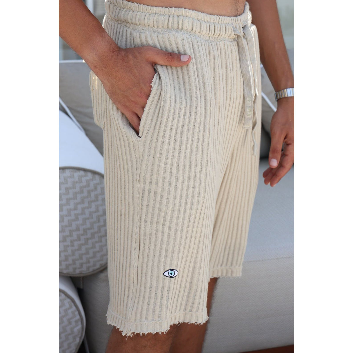 A close-up of a person wearing cream ribbed knit shorts with side pockets and a small blue and white eye logo near the hem, standing indoors near a light-colored sofa.