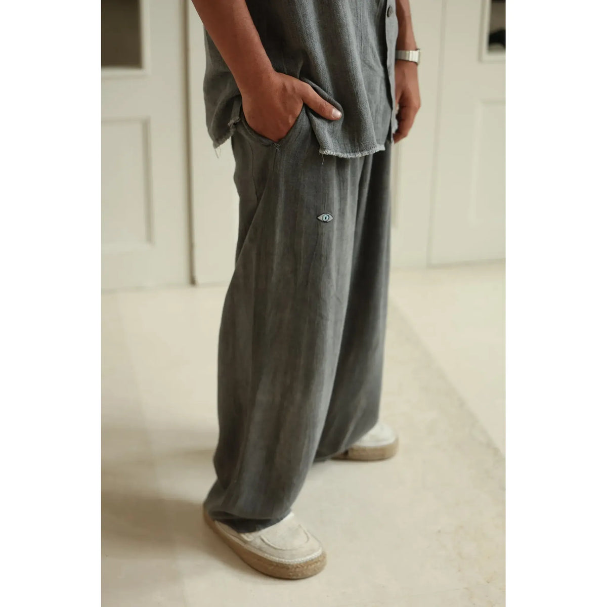 Person wearing Pure Grey Oversized Soft Linen Pants with a small eye emblem and light-colored espadrille shoes