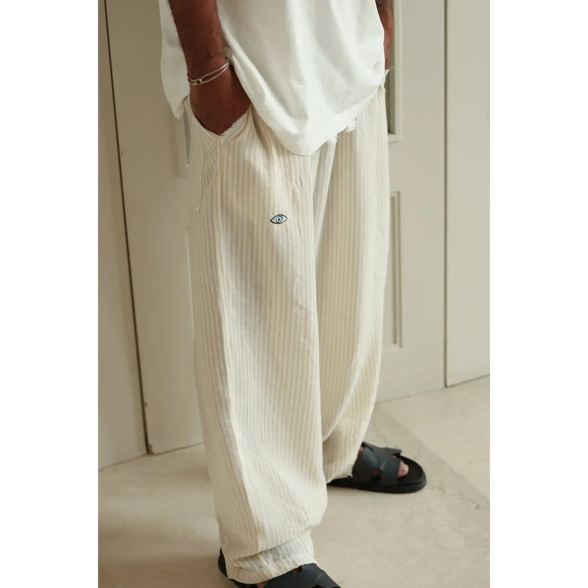 Pure Oversized Linen Pants in yellow and white stripes with a small blue eye logo, worn with black sandals