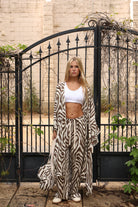 A woman stands in front of a black metal gate wearing a matching beige and black zebra-print robe and wide-leg pants set, styled with a white crop top and white sneakers.