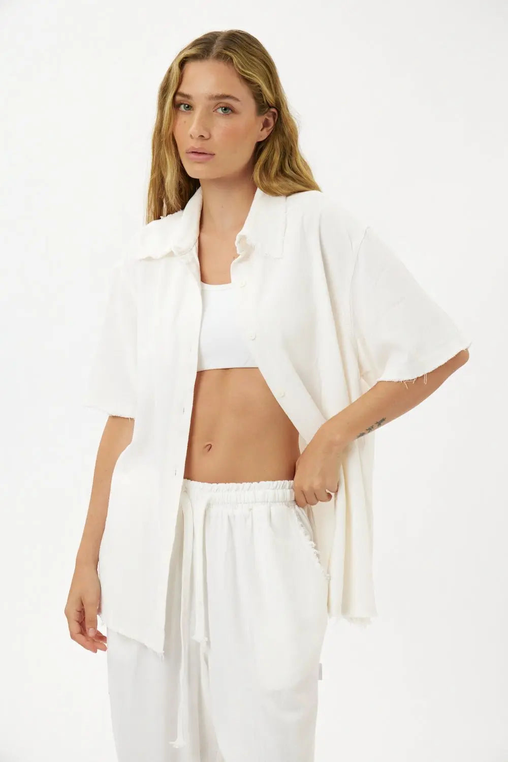 Woman wearing a white Soft Linen Short Sleeve Button-Up Shirt, unbuttoned over a white bralette, and matching white linen pants