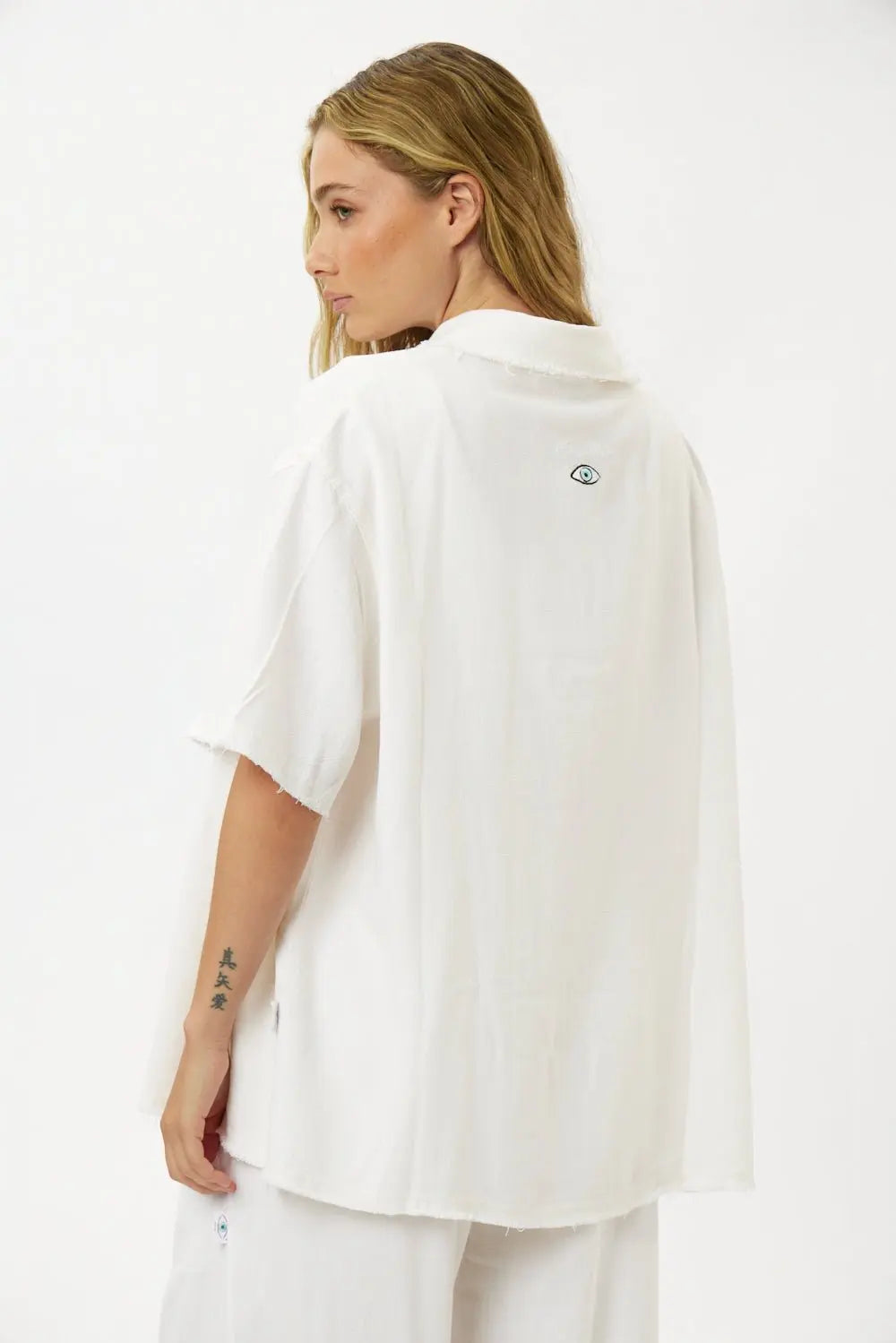 Woman wearing a white Soft Linen Short Sleeve Button-Up Shirt, seen from the back. The shirt has a small eye graphic on the upper back