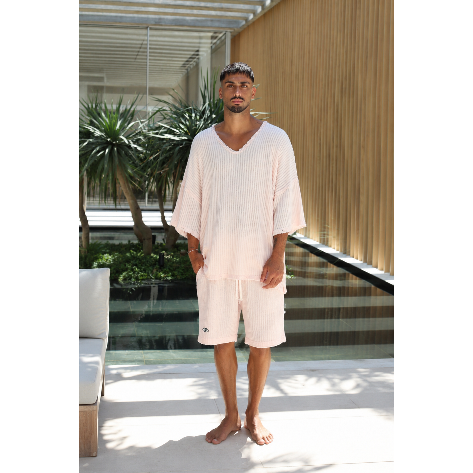 A man wearing a light pink ribbed lounge set consisting of a V-neck tunic top and matching shorts, standing barefoot in a modern outdoor patio setting with plants and a wooden slatted wall.